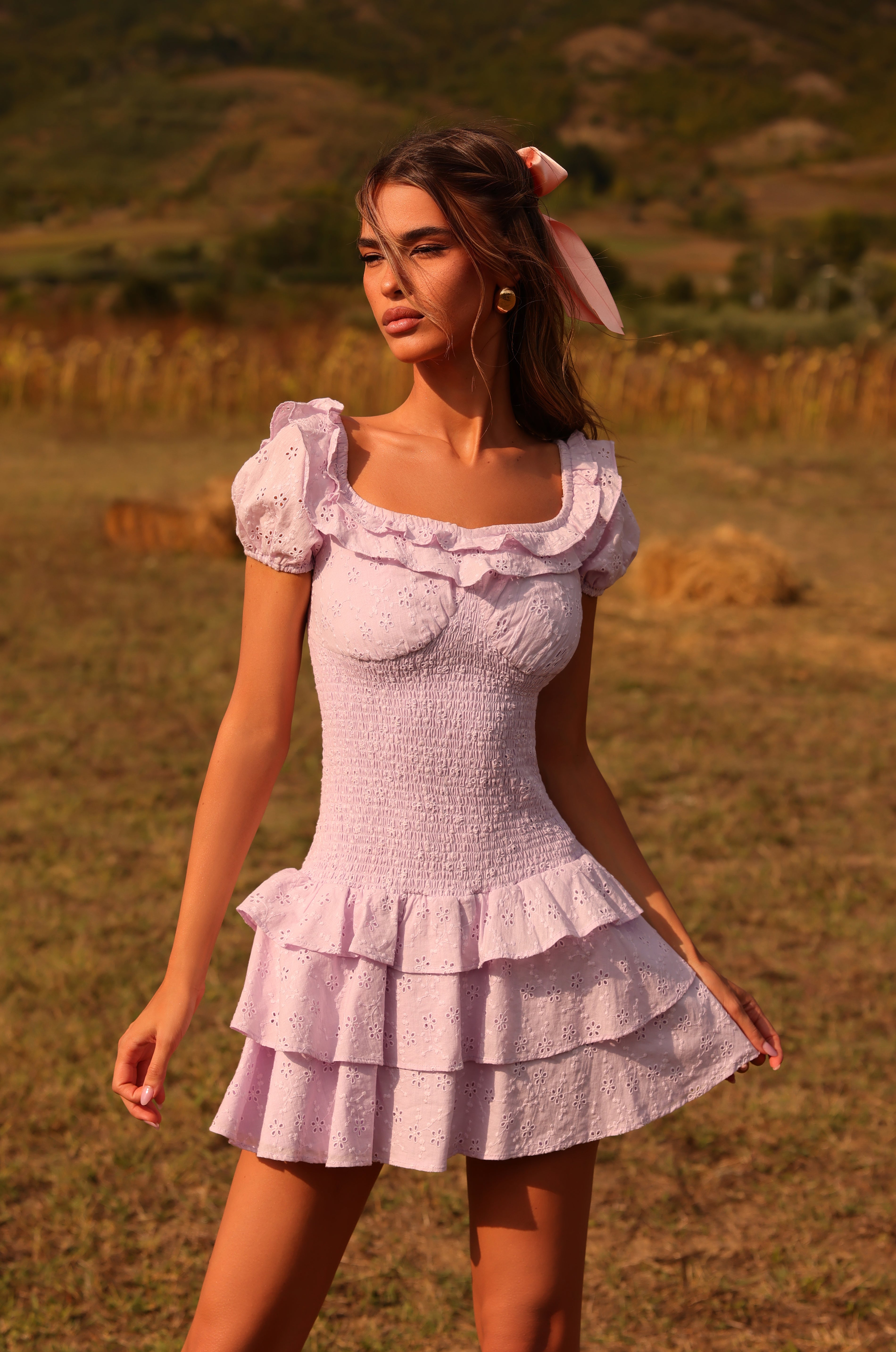 A woman wears the Lilac Purple Short Summer Dress: Macie in light lilac while standing outdoors in a field with mountains in the background under daylight.